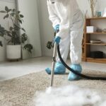 A person in protective gear disinfecting a carpet with a fumigation device indoors.